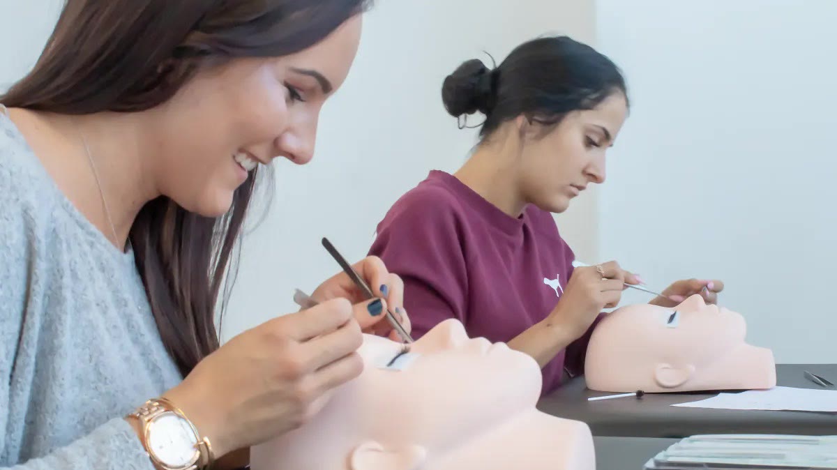 Two lash students practising extension placement on mannequin heads